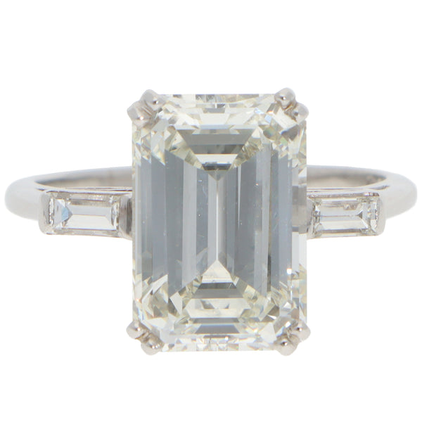 A large emerald cut diamond ring with baguette cut diamond shoulders in white metal, on a white background