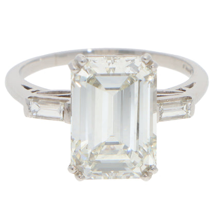 A large emerald cut diamond ring with baguette cut diamond shoulders in white metal, on a white background