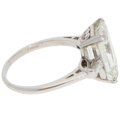 A large emerald cut diamond ring with baguette cut diamond shoulders in white metal, on a white background