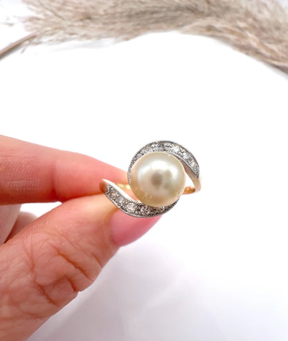 A pearl and diamond crossover swirl ring in yellow and white gold, held in a hand