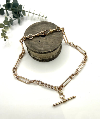 A rose gold trombone and fetter link Albert chain lying on top of a wooden object, on a white background