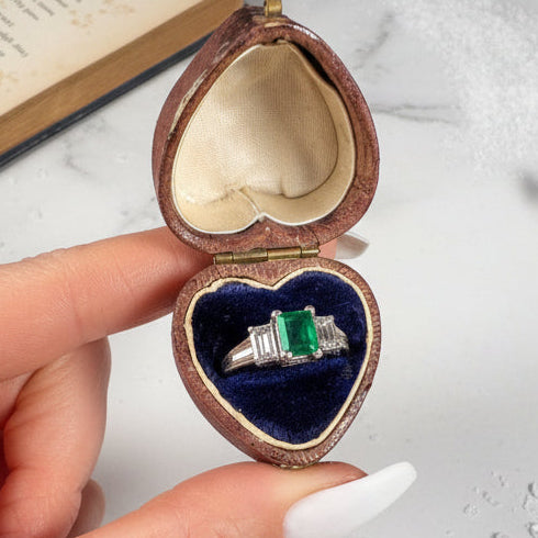 A three stone emerald and diamond ring in platinum, in a heart-shaped antique ring box held by a hand