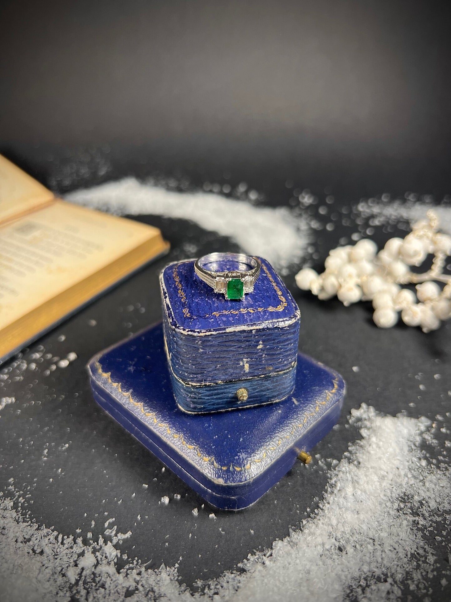 A three stone emerald and diamond ring in platinum, on top of a blue coloured antique ring box