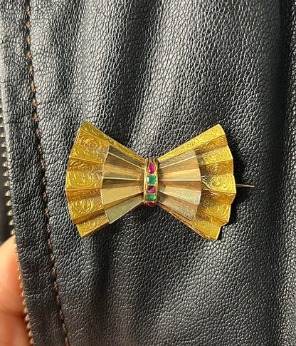 A yellow gold bow shaped brooch with rubies and emeralds, on a black jacket