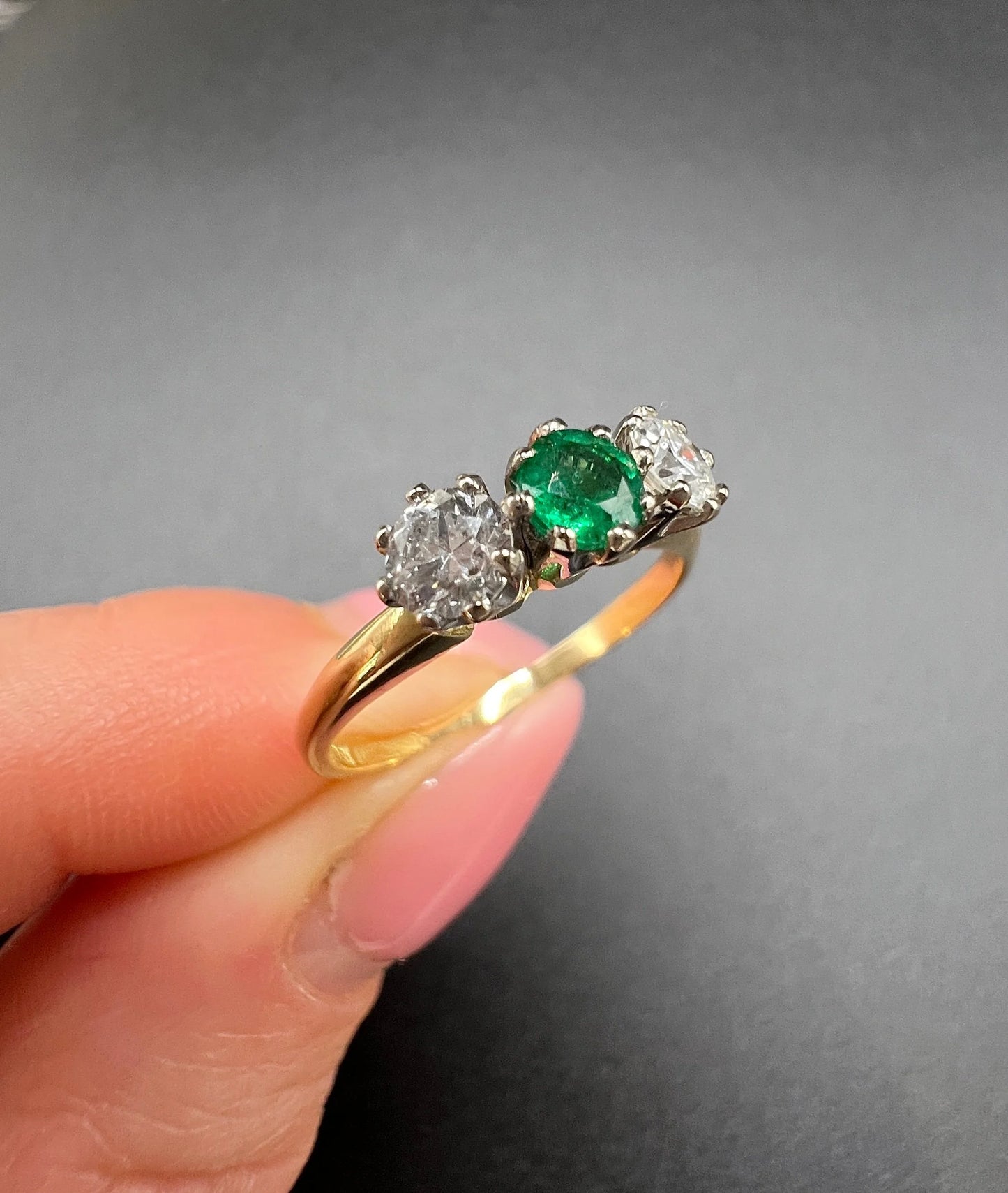 A three stone emerald and diamond ring in yellow gold and platinum, held in a hand