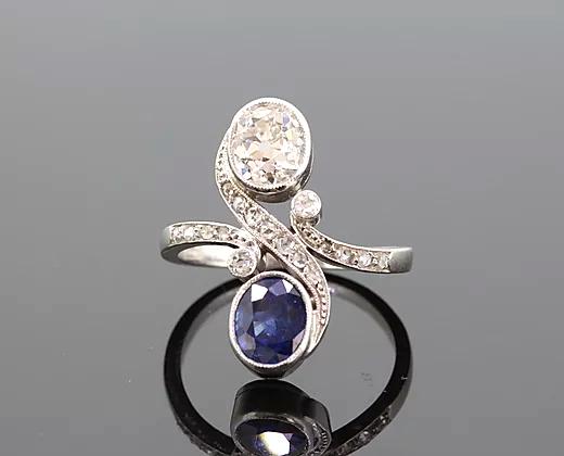 Art Nouveau Sapphire and Diamond Crossover Ring; oval-cut sapphire and oval-cut diamond, accented with diamond set swirls and shoulders, 18ct white gold.