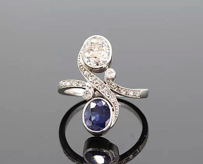 Art Nouveau Sapphire and Diamond Crossover Ring; oval-cut sapphire and oval-cut diamond, accented with diamond set swirls and shoulders, 18ct white gold.