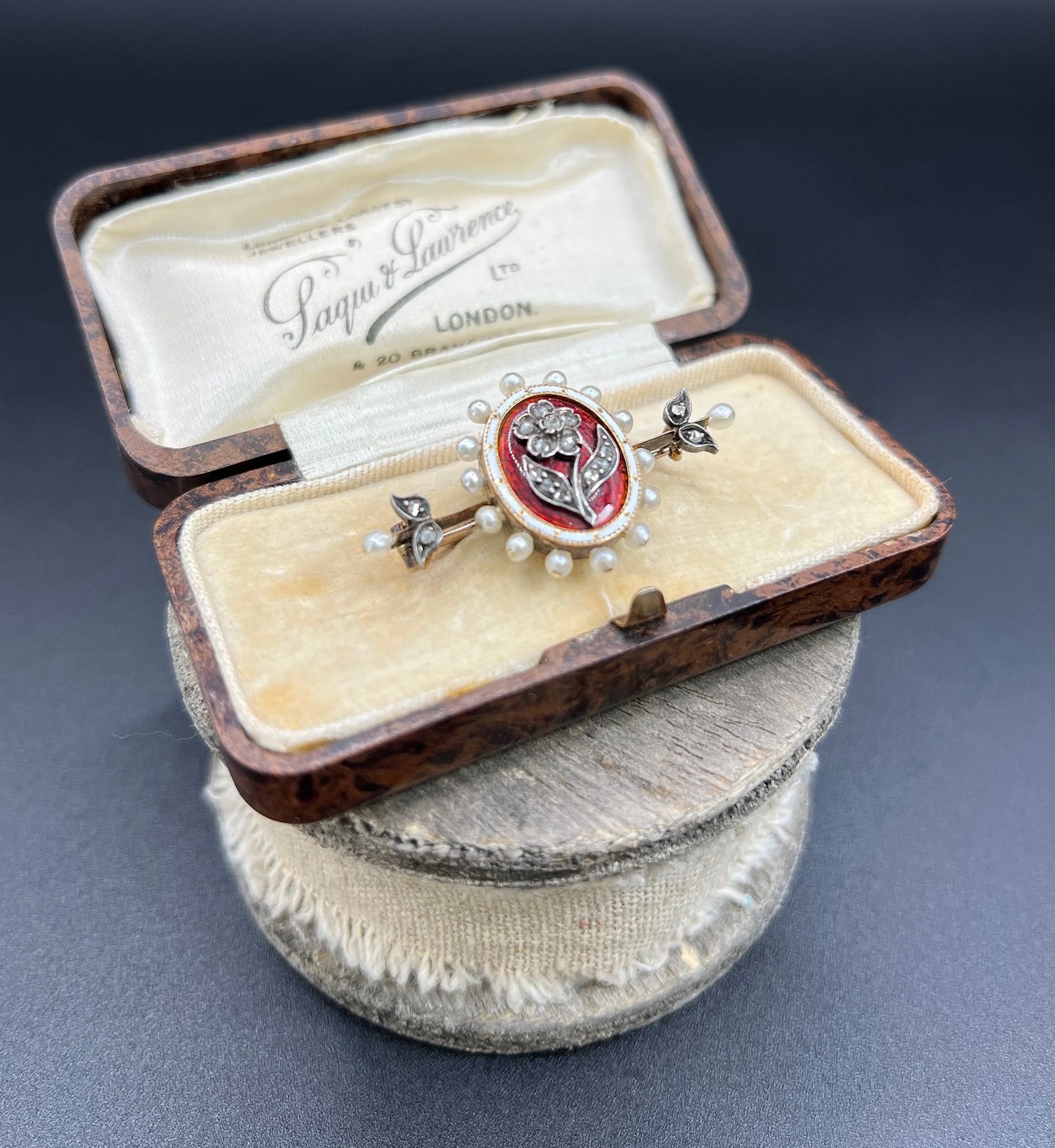 A yellow gold bar brooch with a red enamel centre surrounded by pearls, with a diamond flower motif and diamond leaves at either end, in a box