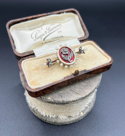 A yellow gold bar brooch with a red enamel centre surrounded by pearls, with a diamond flower motif and diamond leaves at either end, in a box
