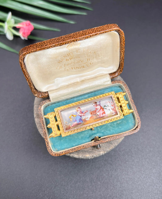 A rectangular shaped yellow gold brooch with an enamelled cherub scene, in an antique box