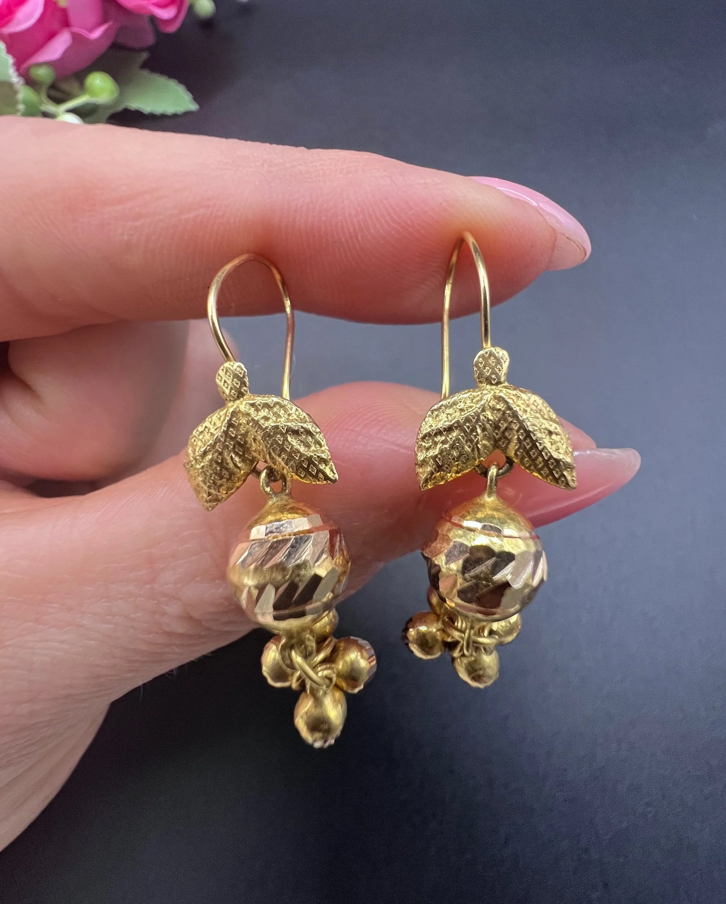 A pair of yellow gold leaf and faceted ball drop earrings with hook fittings, held in a hand
