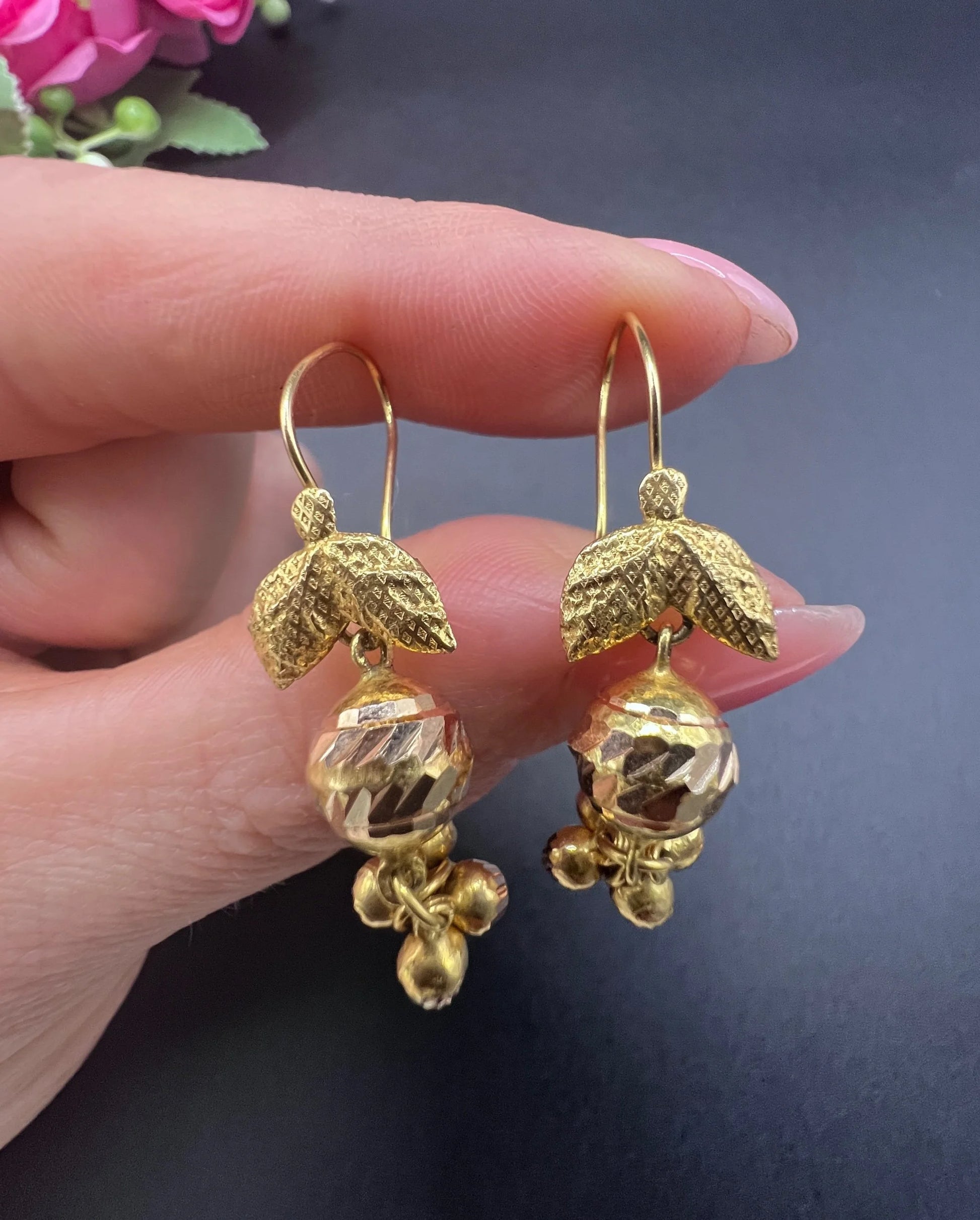 A pair of yellow gold leaf and faceted ball drop earrings with hook fittings, held in a hand