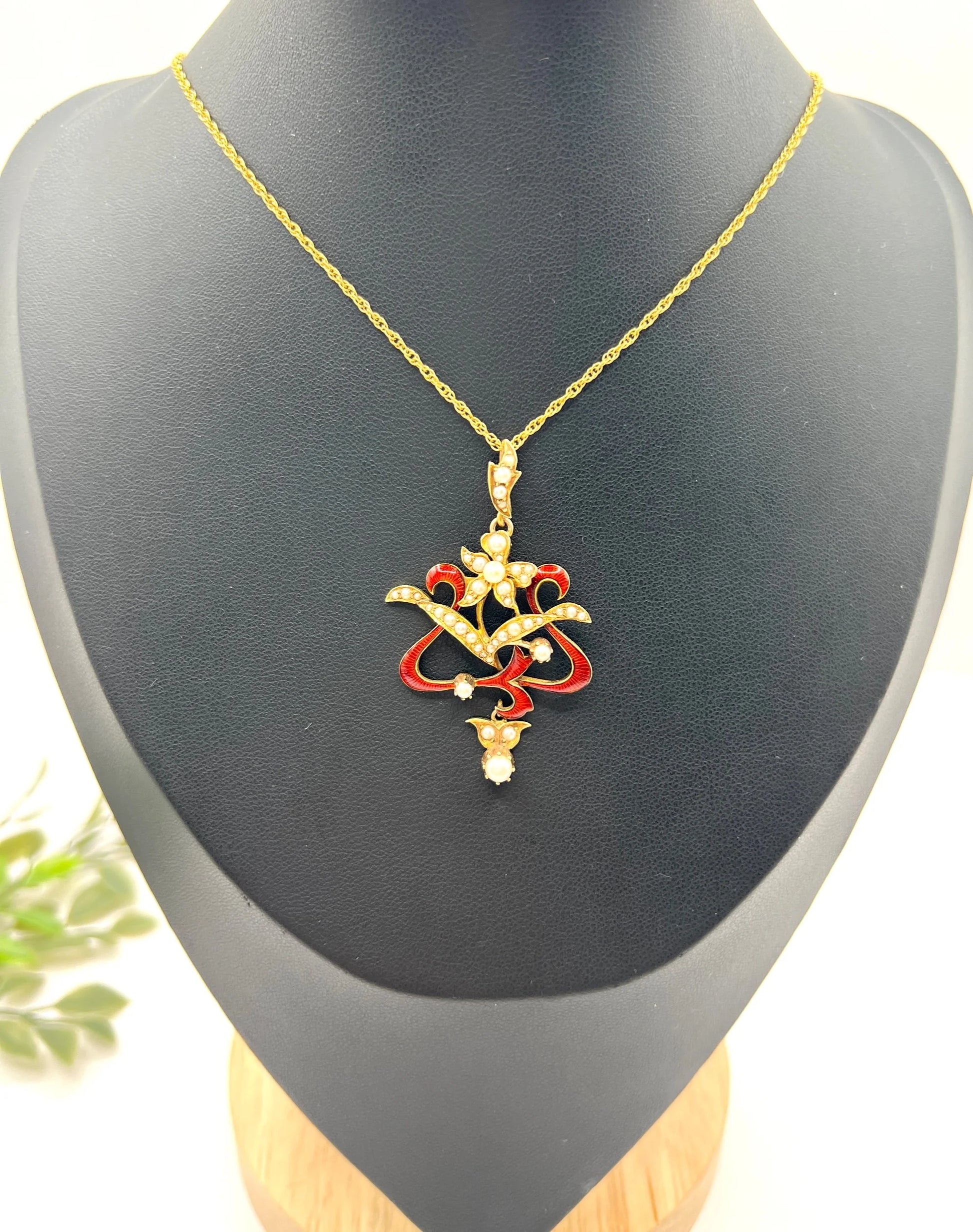 A floral pendant in yellow gold with seed pearls and red enamel, with a chain, on a black jewellery bust