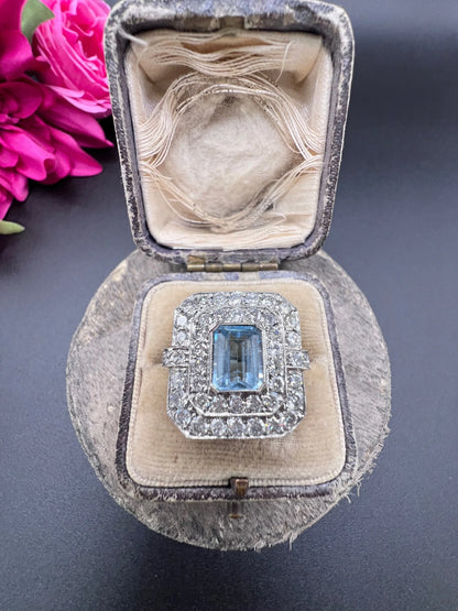 An octagonal shaped cluster ring with an aquamarine centre surrounded by two rows of diamonds, in platinum, in a ring box