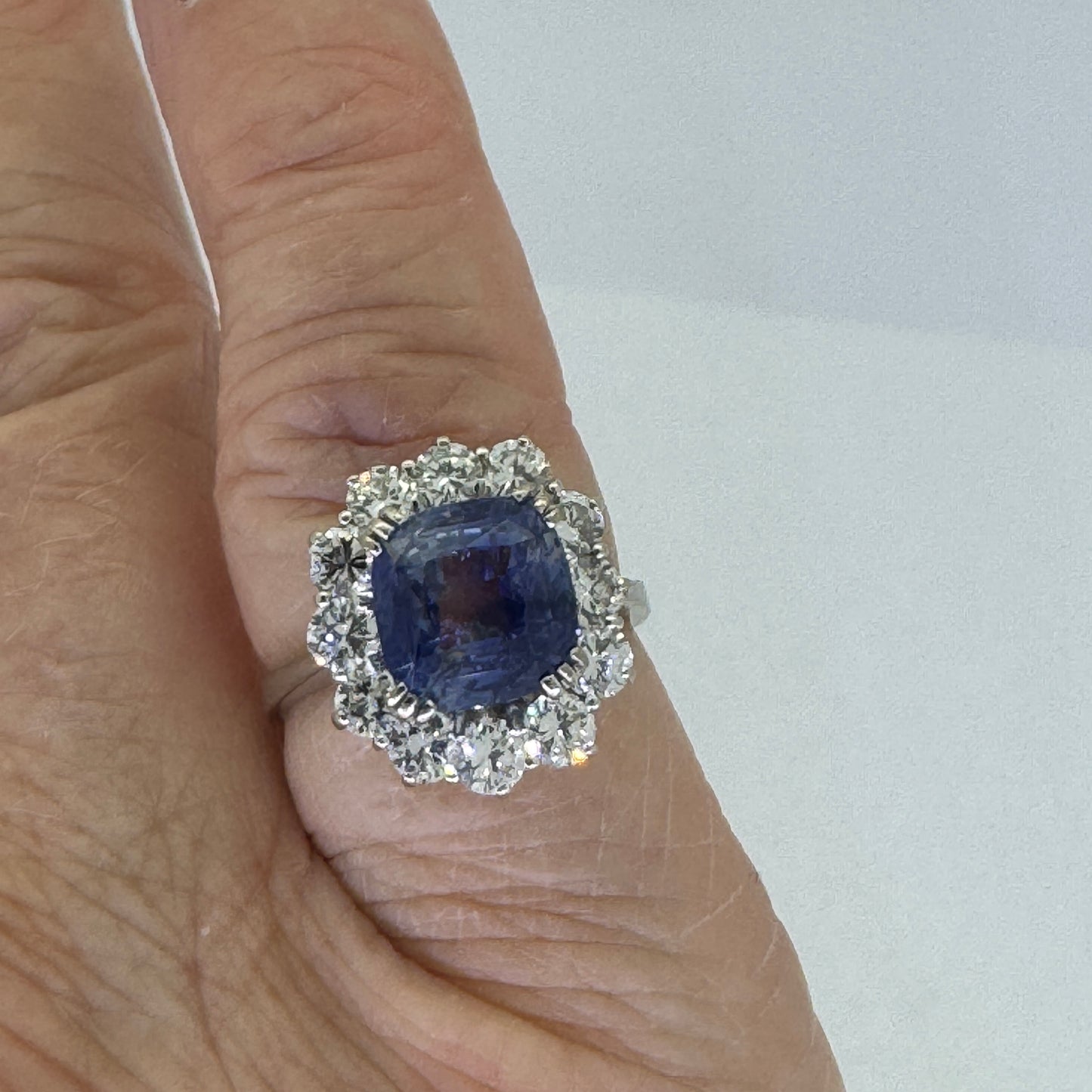 A white gold sapphire and brilliant cut diamond cluster ring modelled on a little finger against a white background
