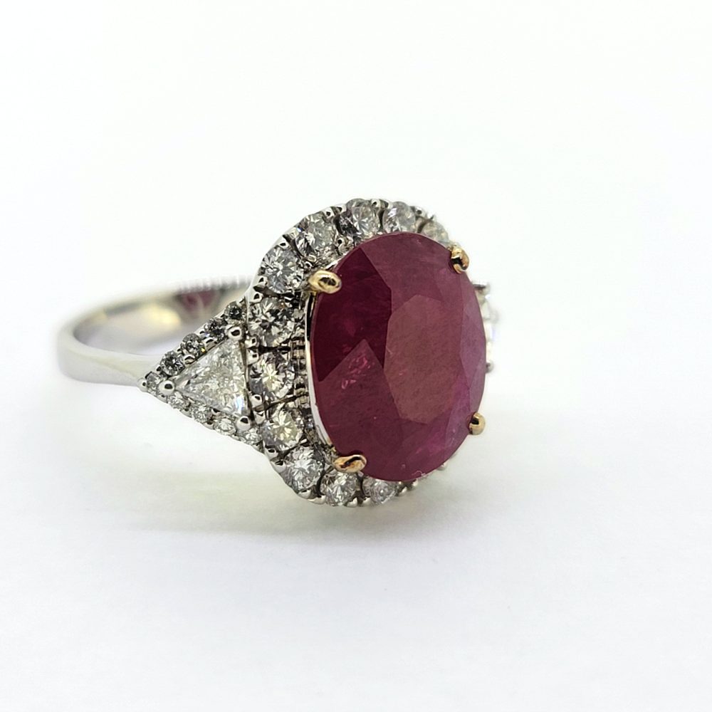Contemporary 4.79ct Ruby and Diamond Cluster Engagement Ring, 4.79ct oval red ruby flanked by trillion diamonds all within a brilliant-cut diamond halo border in 18ct white gold