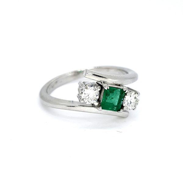 Emerald and Diamond Crossover Three Stone Ring; central princess-cut emerald flanked by brilliant-cut diamonds, in 18ct white gold with stylish crossover design