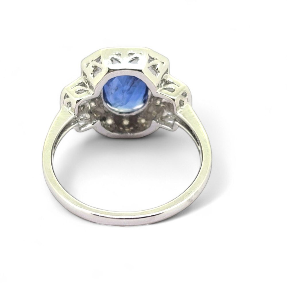 3.3ct Sapphire and Diamond Cluster Dress Ring, 3.30ct oval blue sapphire surrounded by round brilliant-cut diamonds with brilliant and baguette-cut diamond buckle style shoulders in 18ct white gold