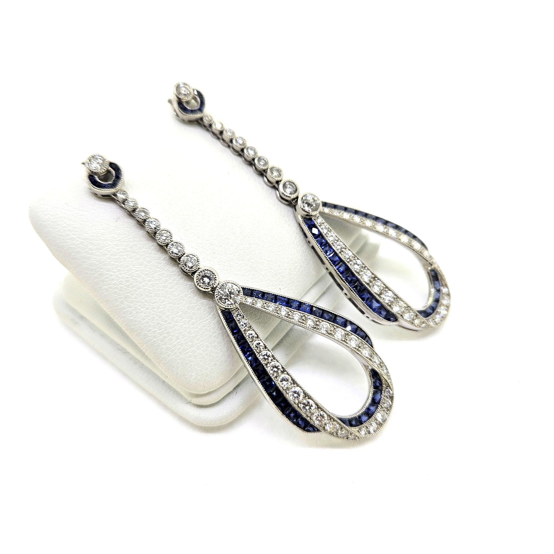 A pair of sapphire and diamond teardrop shaped long drop earrings in white gold, on an earring display pad, against a white background