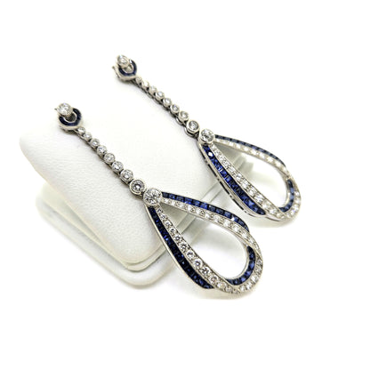 A pair of sapphire and diamond teardrop shaped long drop earrings in white gold, on an earring display pad, against a white background