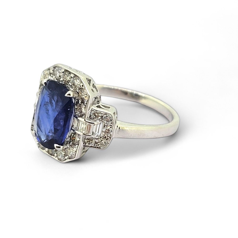 3.3ct Sapphire and Diamond Cluster Dress Ring, 3.30ct oval blue sapphire surrounded by round brilliant-cut diamonds with brilliant and baguette-cut diamond buckle style shoulders in 18ct white gold