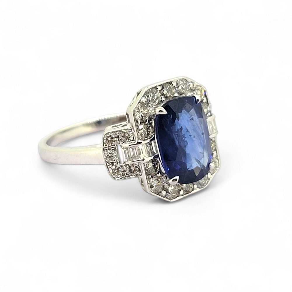 3.3ct Sapphire and Diamond Cluster Dress Ring, 3.30ct oval blue sapphire surrounded by round brilliant-cut diamonds with brilliant and baguette-cut diamond buckle style shoulders in 18ct white gold