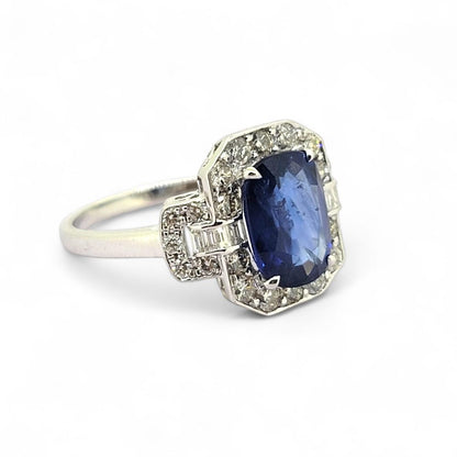 3.3ct Sapphire and Diamond Cluster Dress Ring, 3.30ct oval blue sapphire surrounded by round brilliant-cut diamonds with brilliant and baguette-cut diamond buckle style shoulders in 18ct white gold