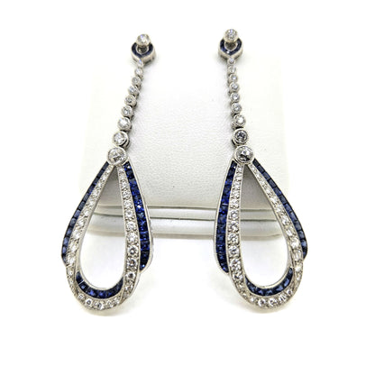 A pair of sapphire and diamond teardrop shaped long drop earrings in white gold, on an earring display pad, against a white background