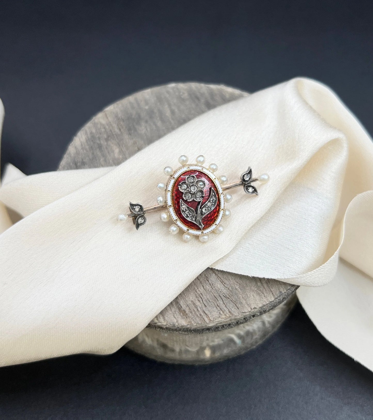 A yellow gold bar brooch with a red enamel centre surrounded by pearls, with a diamond flower motif and diamond leaves at either end