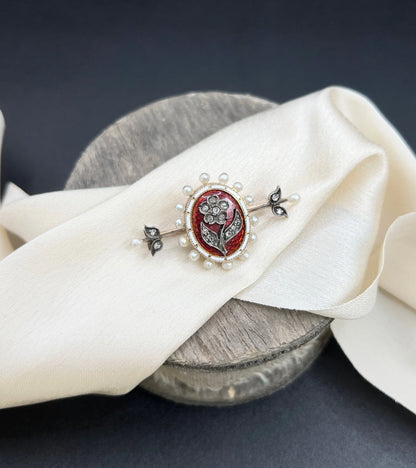 A yellow gold bar brooch with a red enamel centre surrounded by pearls, with a diamond flower motif and diamond leaves at either end