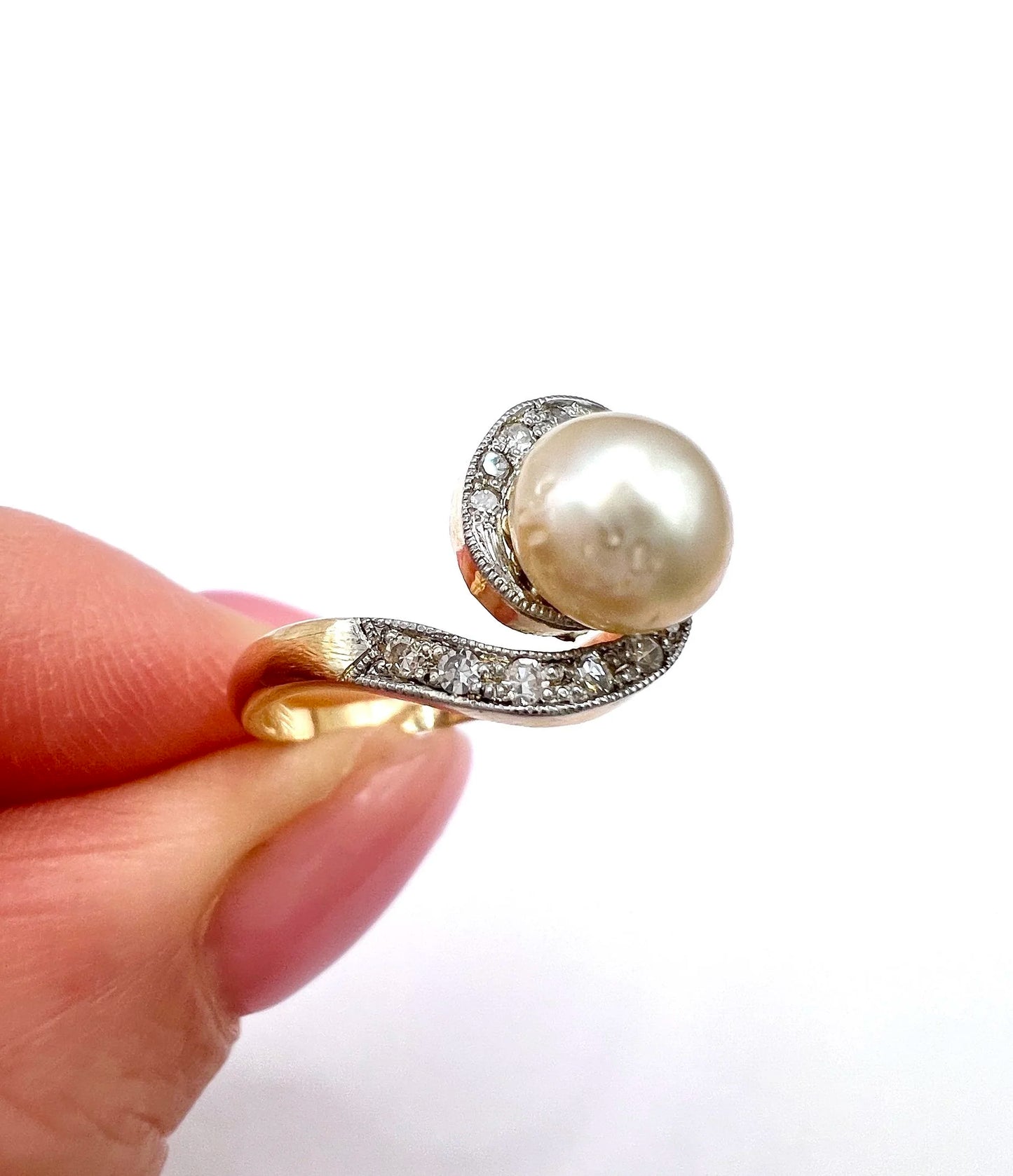 A pearl and diamond crossover swirl ring in yellow and white gold, held in a hand