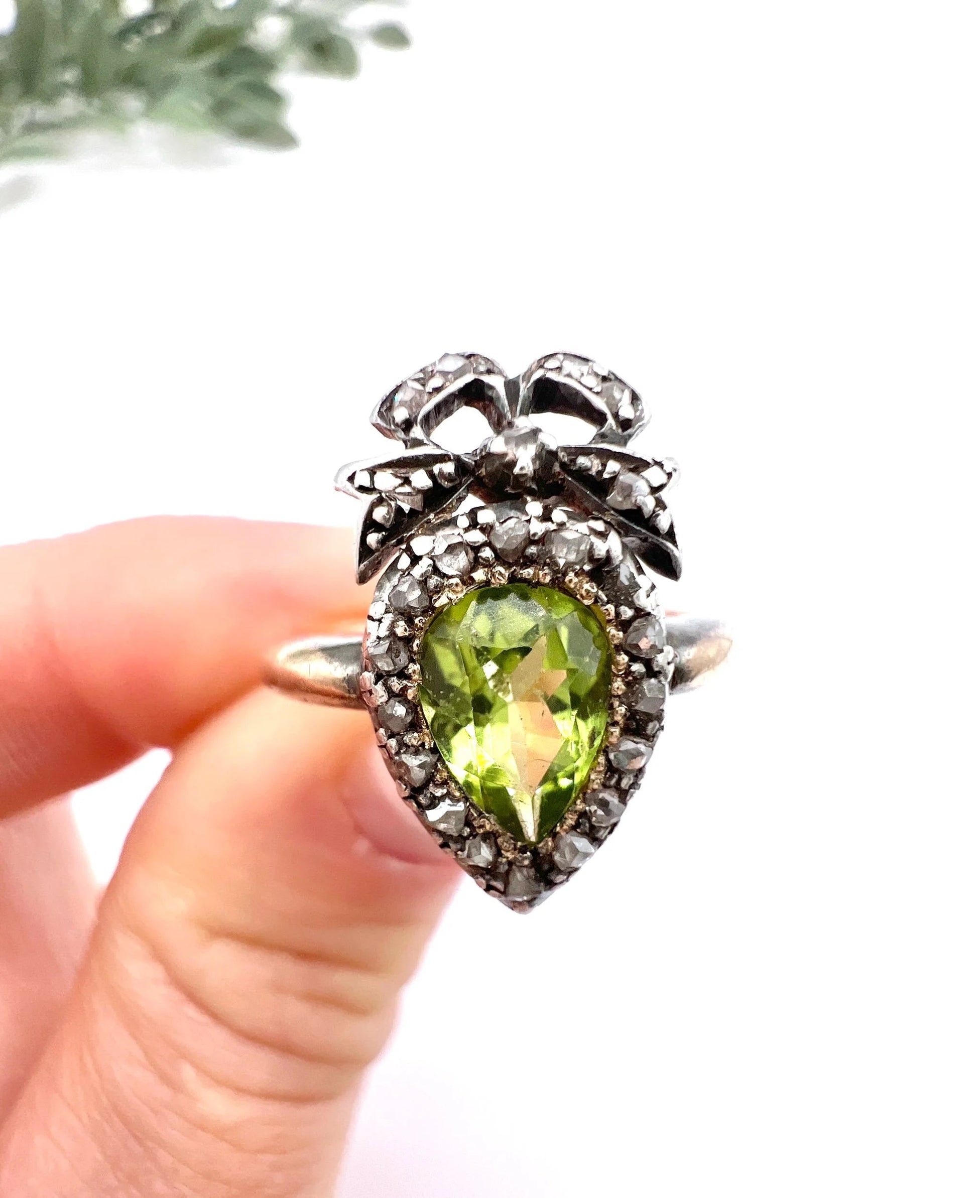 A pear cut peridot and round diamond cluster ring with bow detail, in yellow gold and white metal, held in a hand