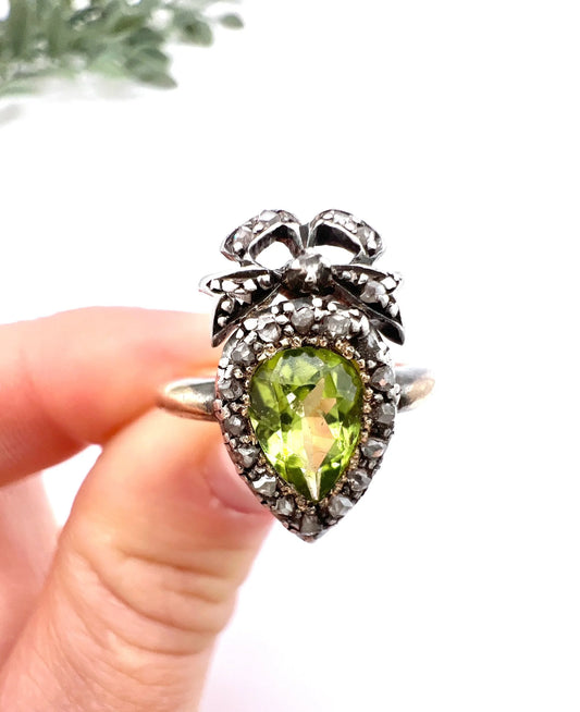 A pear cut peridot and round diamond cluster ring with bow detail, in yellow gold and white metal, held in a hand