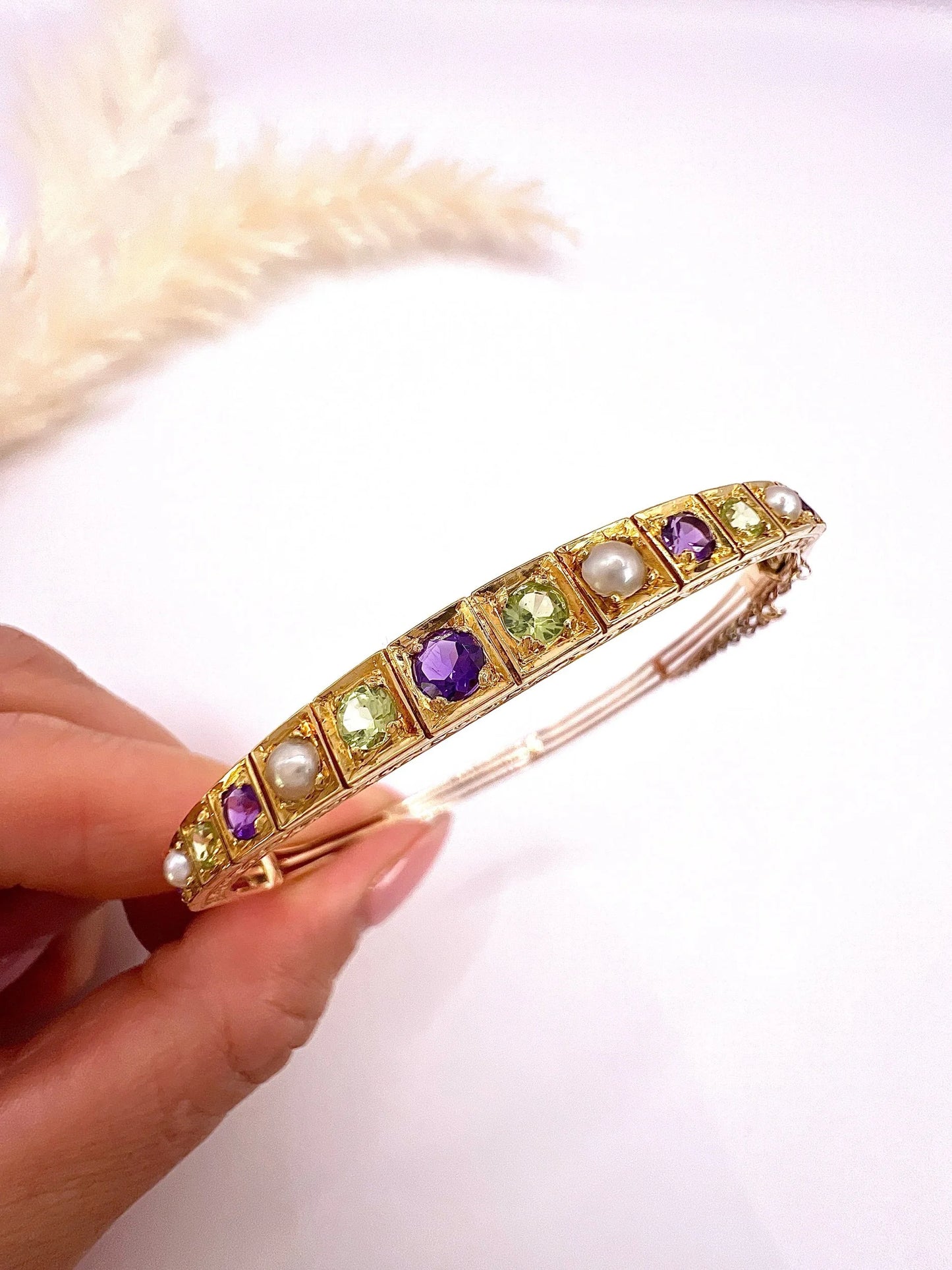 A yellow gold peridot, amethyst and pearl bangle bracelet, held in a hand