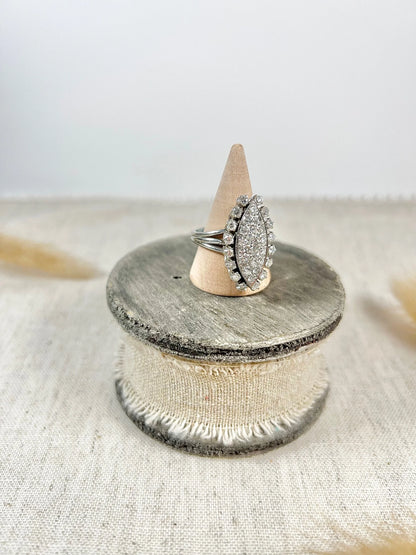 A navette shaped diamond cluster ring in white metal, on a ring cone, sitting on top of a wooden and cloth object
