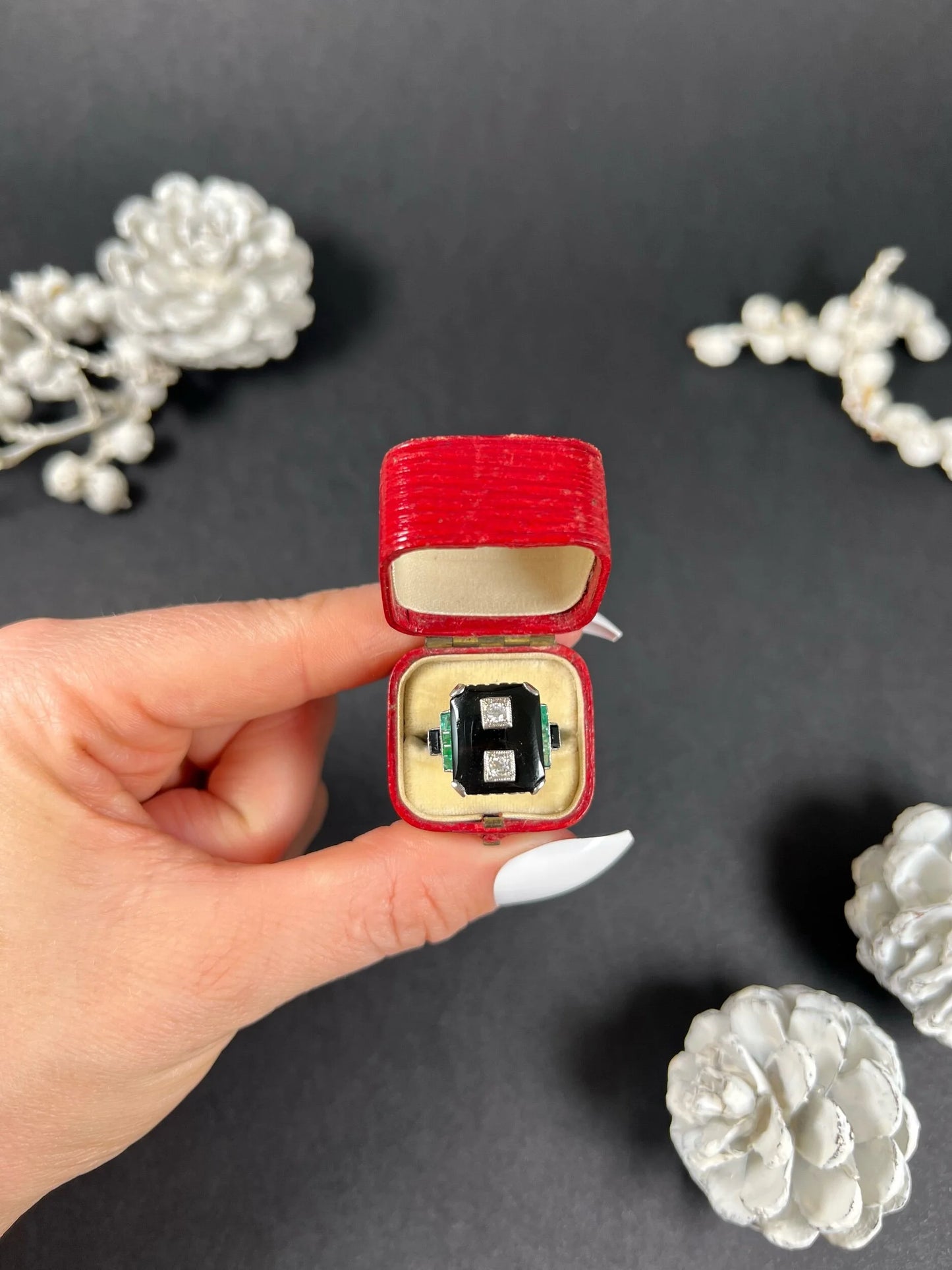 A geometric black onyx ring with diamonds and emeralds in white gold, in an antique ring box held by a hand