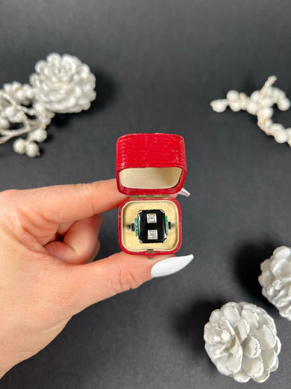 A geometric black onyx ring with diamonds and emeralds in white gold, in an antique ring box held by a hand