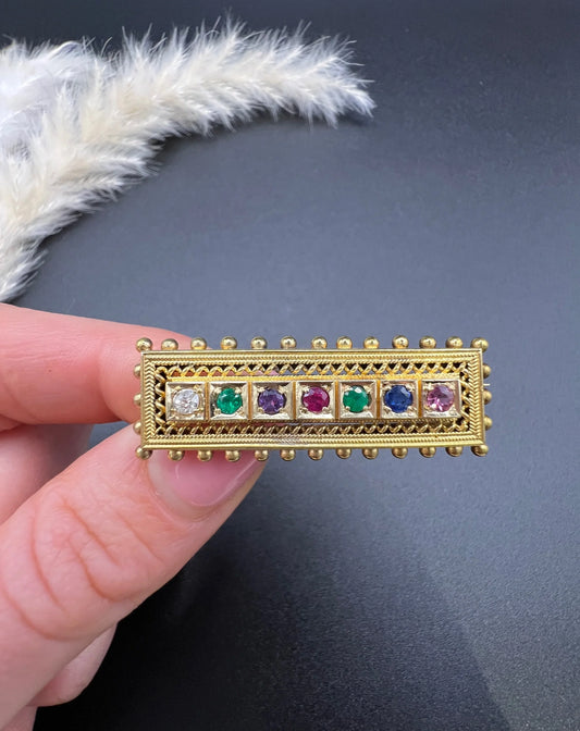 A yellow gold multi-gemstone and diamond acrostic brooch, held in a hand