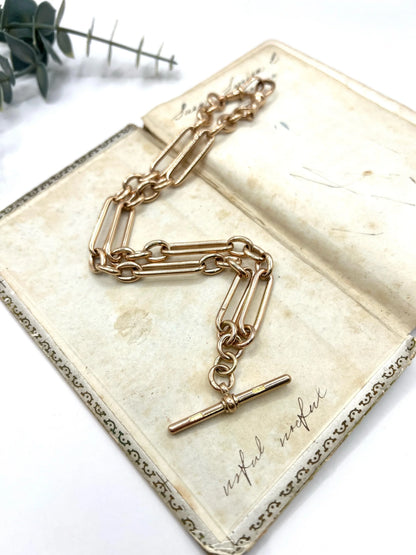 A rose gold trombone and fetter link Albert chain lying on top of an antique book, on a white background