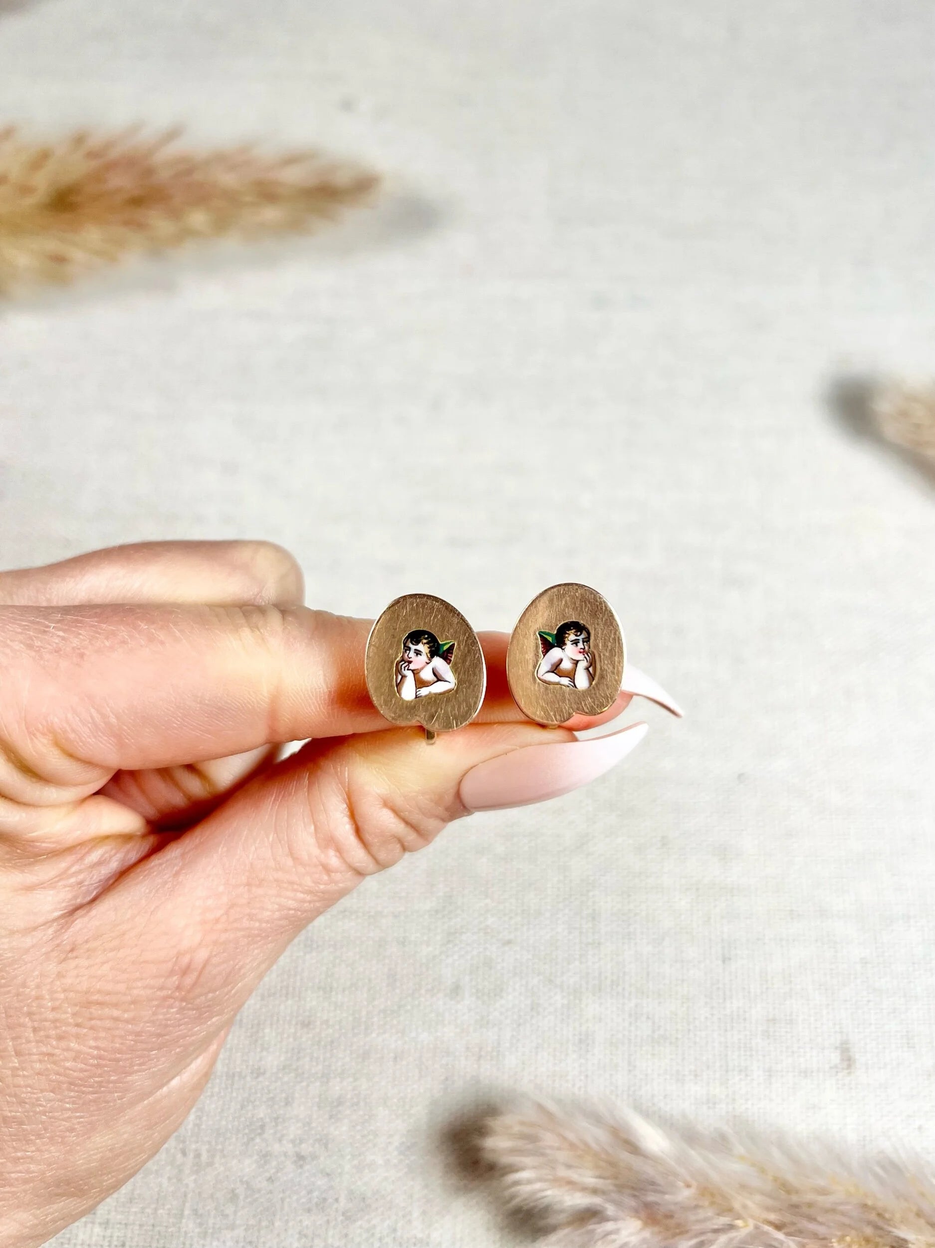 A pair of art palette shaped gold earrings with enamelled cherubs, held in a hand