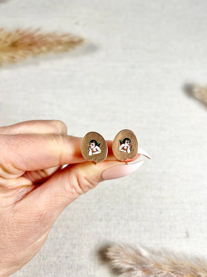 A pair of art palette shaped gold earrings with enamelled cherubs, held in a hand