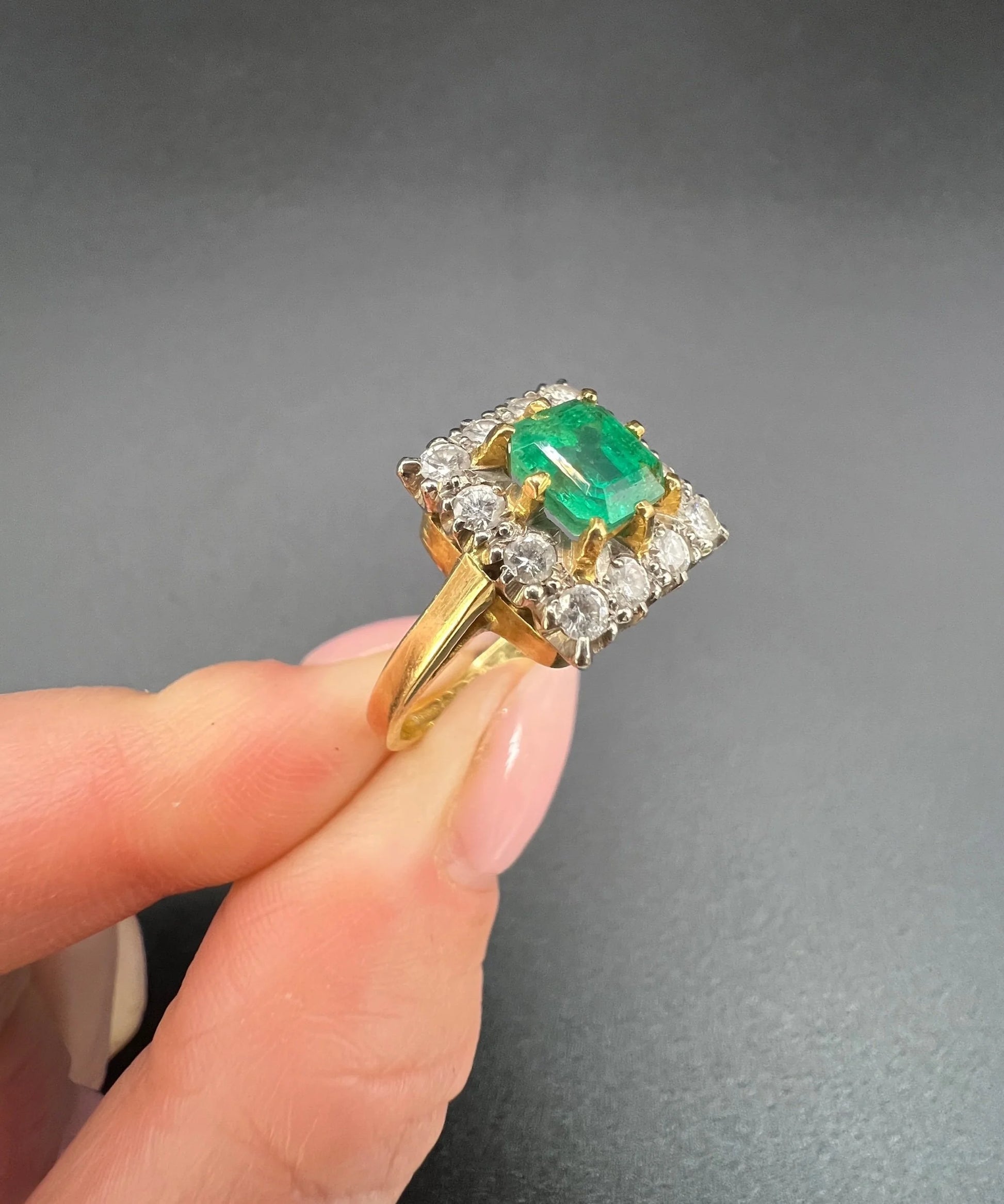 A square shaped emerald and diamond cluster ring in yellow and white gold, held in a hand