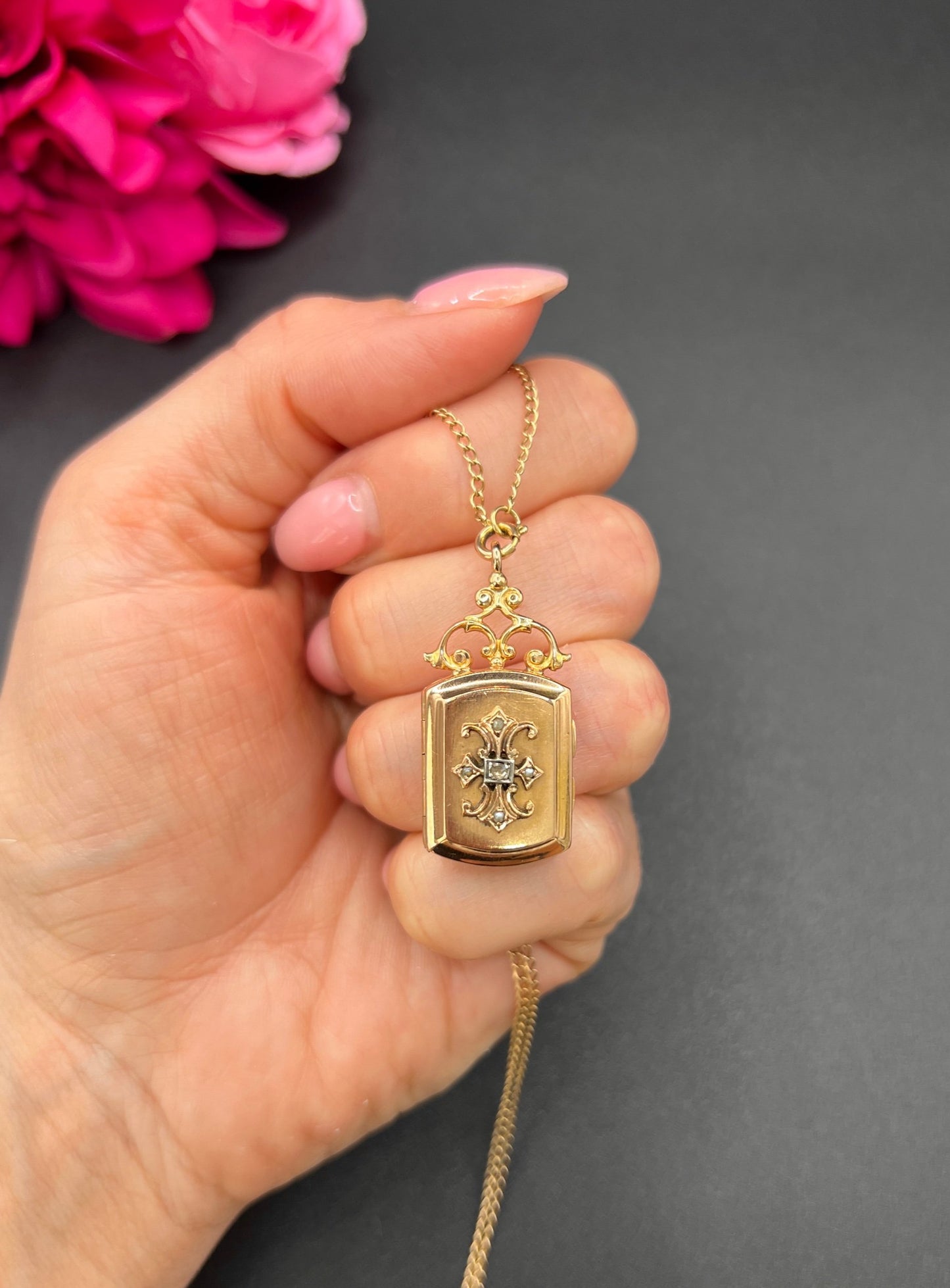 A yellow gold ornate locket with diamond fleur-de-lis detail, with a chain, held in a hand