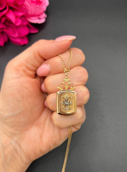 A yellow gold ornate locket with diamond fleur-de-lis detail, with a chain, held in a hand