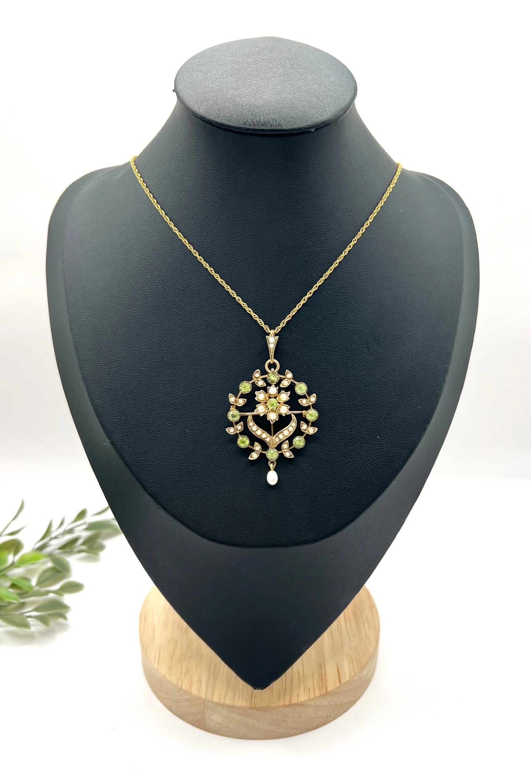 A peridot and pearl floral circle brooch/pendant in yellow gold, with a chain, on a black jewellery bust