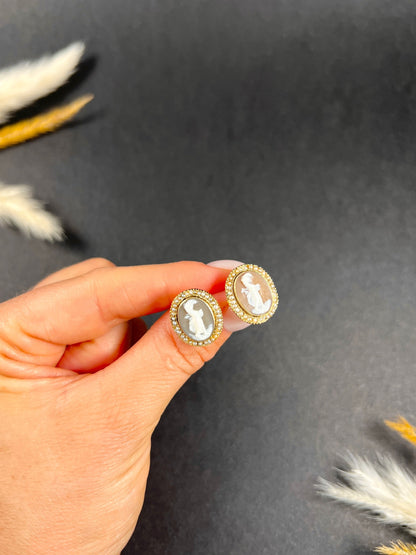 A pair of yellow gold oval shell cameo earrings, held in a hand