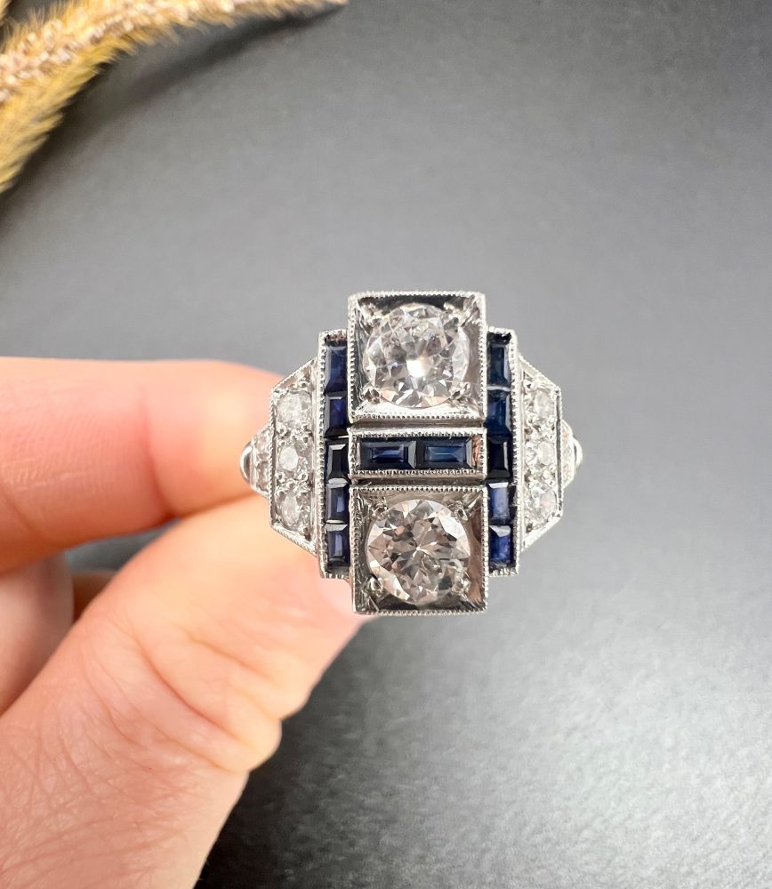 Art Deco Style Sapphire and Diamond Geometric Plaque Ring, two round brilliant cut diamonds in square bezel settings bordered by calibre sapphires in 18ct white gold with diamond stepped shoulders