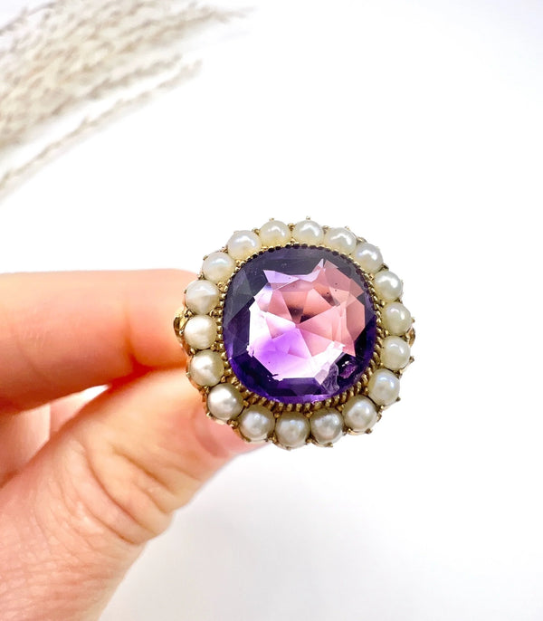 A cushion shaped amethyst cluster ring with a pearl border, in yellow gold, held in a hand