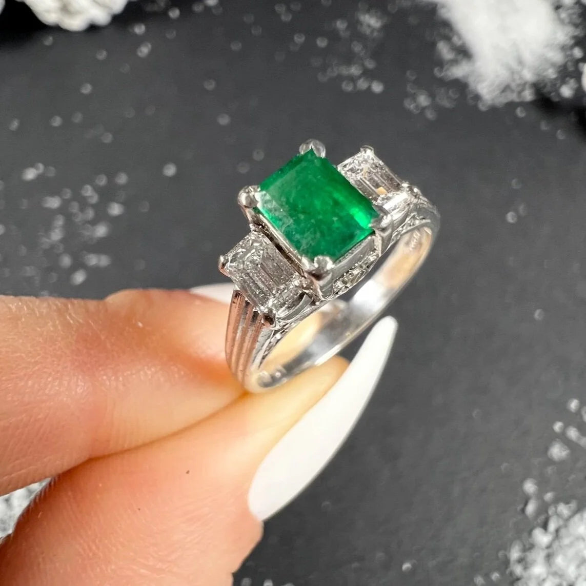 A three stone emerald and diamond ring in platinum, held in a hand