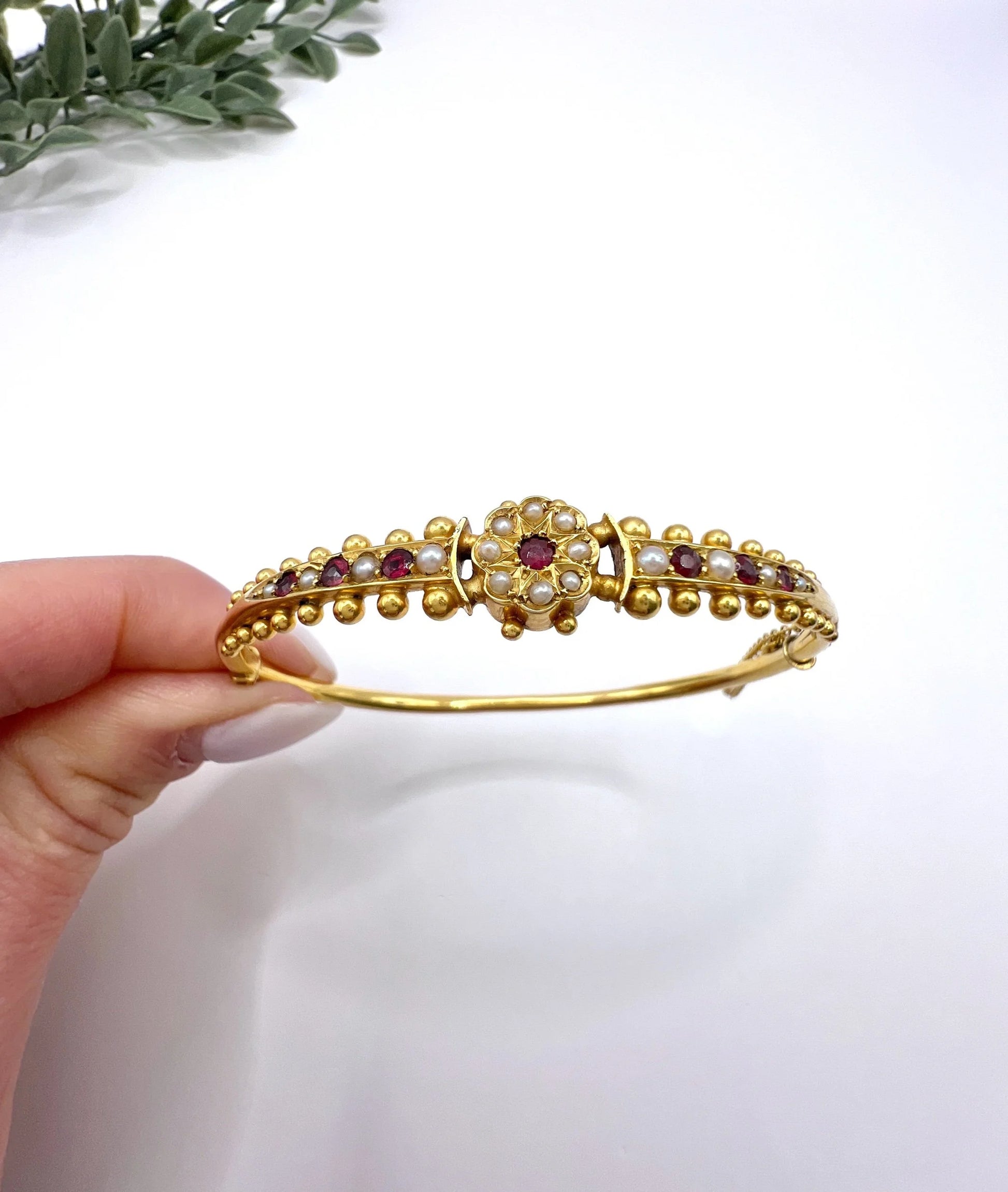 A yellow gold bangle bracelet set with rubies and seed pearls, held in a hand